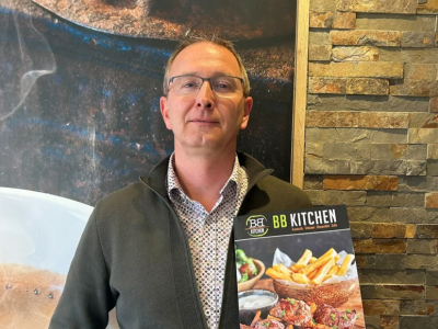 Man toont promotiefolder van BB Kitchen met gerechten op de voorgrond tegen een steenmuur achtergrond.