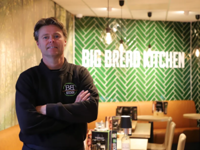 Man in BB Kitchen-uniform staat met gekruiste armen in een sfeervolle eetgelegenheid met groen betegelde muur.