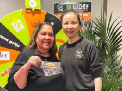 Twee medewerkers van BB Kitchen staan voor een rad van fortuin met prijzen en een BB Kitchen-poster op de achtergrond.