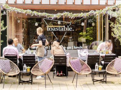 Buiten terras van Toastable met klanten etend en zittend onder een groene guirlande voor de glazen gevel met logo.