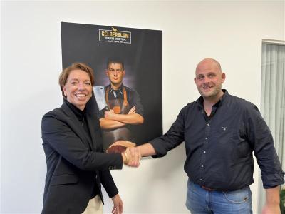 Twee mensen schudden elkaar de hand voor een Gelderblom-posters, waarbij de samenwerking benadrukt wordt.