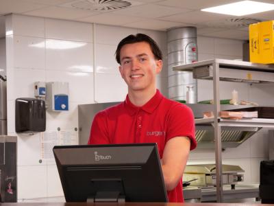 Medewerker in rood uniform bij kassa in een moderne keuken van een fastfoodrestaurant.