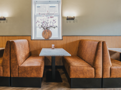 Brasserie-inrichting met bruine leren zitbanken, marmeren tafels en een vaas met bloemen voor het raam.