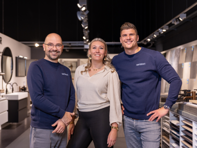 Drie mensen in een Sanidirect-showroom, gekleed in bedrijfskleding, poseren lachend voor de camera.