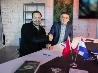 Twee mannen schudden handen over een tafel met Turkse en Nederlandse vlaggetjes en documenten.
