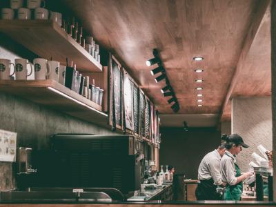 Twee barista's werken achter de counter van een koffiebar met menu’s en kopjes aan de muur.