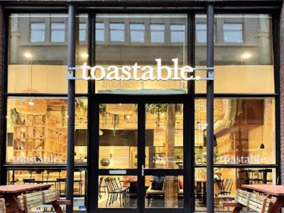 Gevel van een Toastable-restaurant in Den Haag met grote ramen en uitnodigende houten tafels buiten.