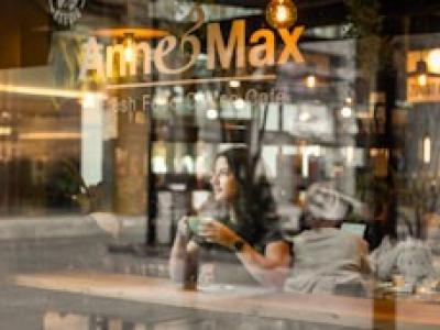 Een vrouw geniet van koffie in een sfeervolle Anne&Max-vestiging met het logo zichtbaar op het raam.