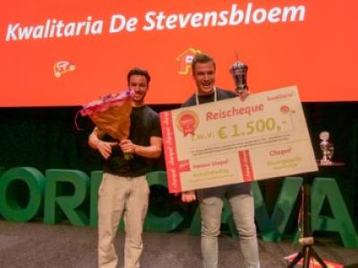Twee lachende mannen op een podium met een grote cheque en bloemen, onder de tekst Kwalitaria De Stevensbloem.
