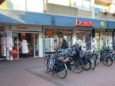Bruna-winkel in een winkelstraat, met mensen en geparkeerde fietsen ervoor.