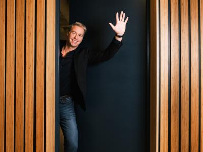 Man in een deuropening van een houten kantoorruimte, gekleed in een donker jasje, zwaait met een glimlach.