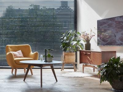 Gezellige woonkamer met gele fauteuil, salontafel, groene planten, houten dressoir en muur met jaloezieën.