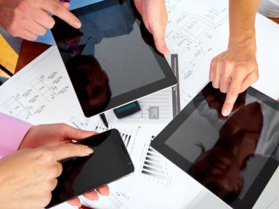 Mensen wijzen naar tablets en smartphone bovenop technische tekeningen en grafieken op een bureau.