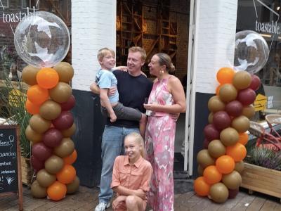 Een gezin poseert lachend voor de ingang van hun Toastable-vestiging met kleurrijke ballonpilaren.