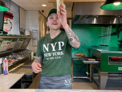 Medewerker van New York Pizza draait pizzadeeg in de lucht achter de balie met gereedschappen en menu op de achtergrond.