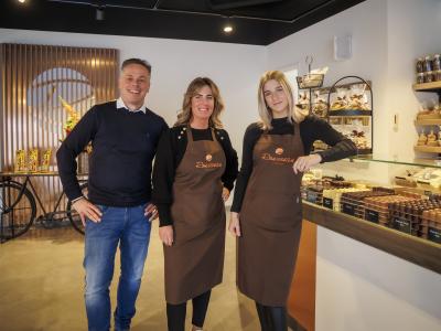 Drie medewerkers poseren lachend in een charmante chocoladewinkel met uitgestalde lekkernijen.