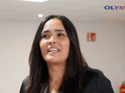 Vrouw met donker haar spreekt enthousiast in een kantoorruimte met Olympia-logo op de achtergrond.