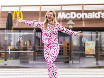 Vrouw in roze-wit pak met uitgestrekte armen voor een McDonald's restaurant.