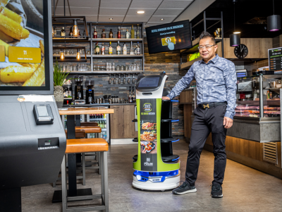Man poseert naast een bezorgrobot in een restaurantinterieur met bestelzuil en bar op de achtergrond.