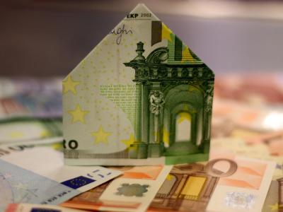 Een huisvormige constructie gevouwen van een biljet van 100 euro, geplaatst op een stapel van andere eurobiljetten. De afbeelding symboliseert de kosten van huisvesting of vastgoed in geldtermen.