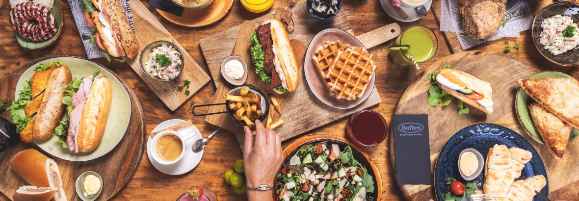 Een uitgebreide variatie aan Bufkes-producten op een houten tafel, waaronder broodjes, soepen, salades en drankjes.