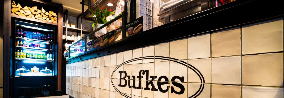 Toonbank met Bufkes-logo, koelkasten met dranken en etenswaren en houten decoratie in een fraai ingerichte zaak.