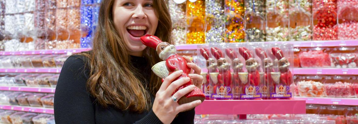Vrouw in een Jamin-winkel houdt een chocolade Sinterklaas vast met kleurrijke snoepachtergrond.