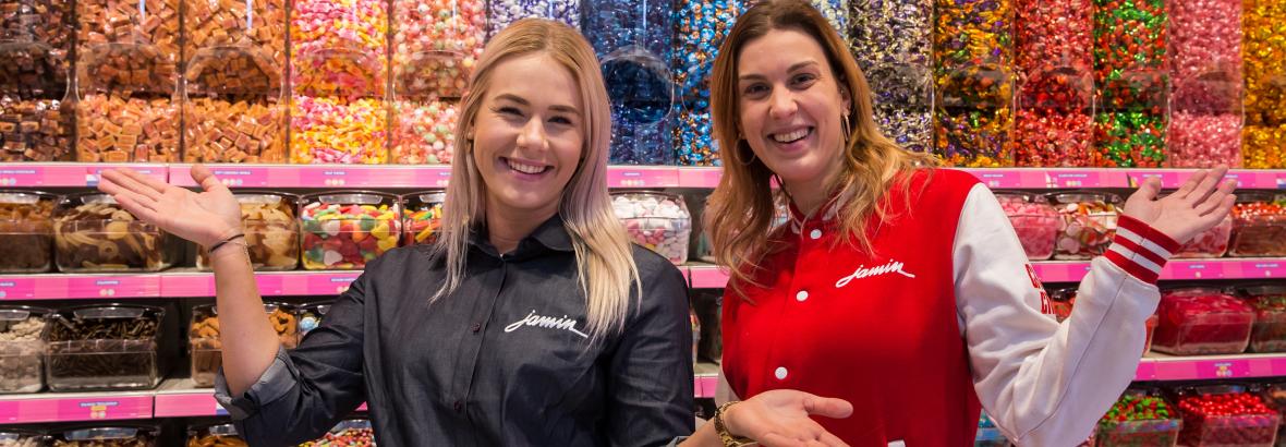 Twee medewerkers bij een kleurrijke snoepwand in een Jamin-winkel, omgeven door diverse snoepvariaties.