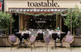 Terras van Toastable Arnhem met tafels, stoelen en decoratieve kussens, omgeven door groene planten.
