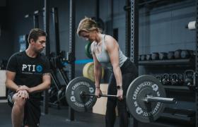 Vrouw doet gewichthefoefening in sportschool terwijl trainer toekijkt.