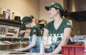 Medewerkers van New York Pizza bereiden pizza's voor in een moderne keuken, gekleed in groene bedrijfskleding.