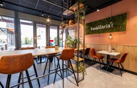 Interieur van een Kwalitaria met houten tafels, bruine stoelen, groene planten en een moswand met het logo.