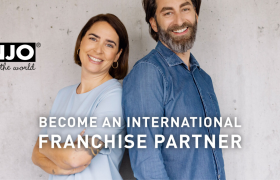 Twee lachende mensen staan rug aan rug voor een betonnen muur met de tekst Become an International Franchise Partner.