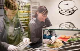 Medewerkers bereiden sandwiches in een BackWERK!-winkel achter een toonbank met vers beleg en broodjes.