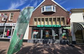 Voorgevel van een New York Pizza vestiging in een bakstenen pand met een groene vlag en diverse reclameposters op de ramen.