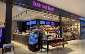 Vooraanzicht van een Bakker Bart winkel met broodschappen, koelvitrines en digitale menuschermen onder een verlicht roze logo.