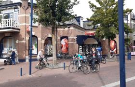 Exterieur van een bakstenen hoekpand met een Bruna-winkel, fietsen op de stoep en voorbijgangers op straat.