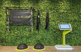 Trainingsstation met Wav-e Studios logo tegen een groene plantenwand, voorzien van fitnessattributen en een wit-groen bedieningspaneel.