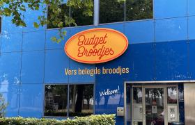 Een blauwe gevel met het Budget Broodjes logo, de tekst Vers belegde broodjes en een glazen toegangsdeur met huisnummer 77.