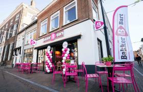 Exterieur van een Bakker Bart op een straathoek met een terras van roze stoelen, witte en roze ballonpilaren en een reclamevlag aan de gevel.