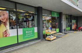 Voorgevel van een Read Shop winkel met grote glazen ruiten, een groen logo, een tijdschriftenrek buiten en een rode scooter op de stoep.