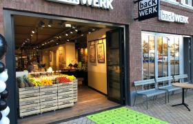 De entree van een backWERK vestiging met houten kratten vol vers fruit bij de ingang, een groen tapijt en buitenmeubilair tegen de gevel.