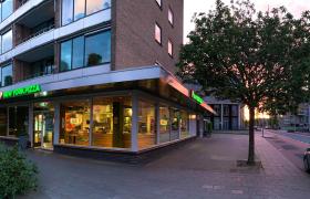 Buitenaanzicht van New York Pizza op een hoek onder een appartementencomplex bij zonsondergang, met glazen puien en verlichte logo's.